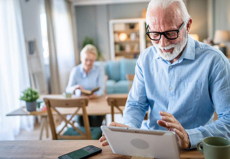 Älterer Mann am Tablet im Wohnzimmer, Frau im Hintergrund